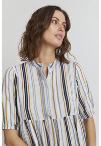 Blusa a righe con bottoni in tessuto leggero. I colori includono bianco, blu navy, oro e grigio. Presenta un colletto a bande e una parte inferiore raccolta e fluida.