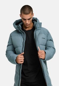Padded jacket in light blue, featuring a hood, front zipper, and black logo detail. Worn over a black shirt, showcasing smooth texture.
