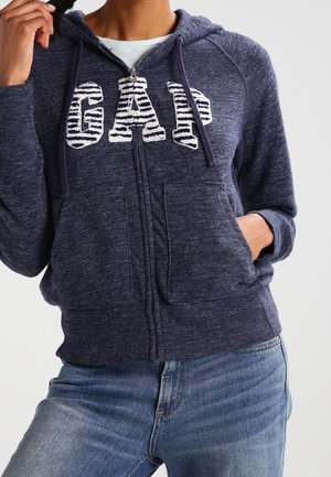Frau trägt einen marineblauen Kapuzenpullover mit Reißverschluss mit "GAP" in weißen gestreiften Buchstaben und hellblauen Jeans, eine Hand in der Tasche und die andere hält die Kordel.
