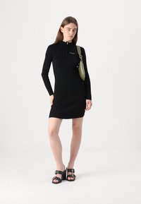 Črna rebrasta obleka z dolgimi rokavi, ki ima zadrgo na vratu in zelene detajle. Model nosi zeleno torbico in črne sandale.