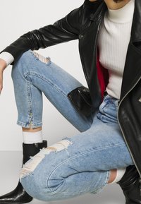 Vaqueros de denim azul rasgados con un acabado desgastado, cortos a la altura del tobillo. Combinados con una chaqueta de cuero negra y una camiseta blanca de canalé.