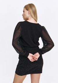 Schwarzes Oberteil mit durchsichtigen, gepufften Ärmeln mit silbernen Punkten. Kombiniert mit einem schwarzen Rock mit weißen Polka-Dots. Die Hände des Modells sind hinter dem Rücken positioniert.