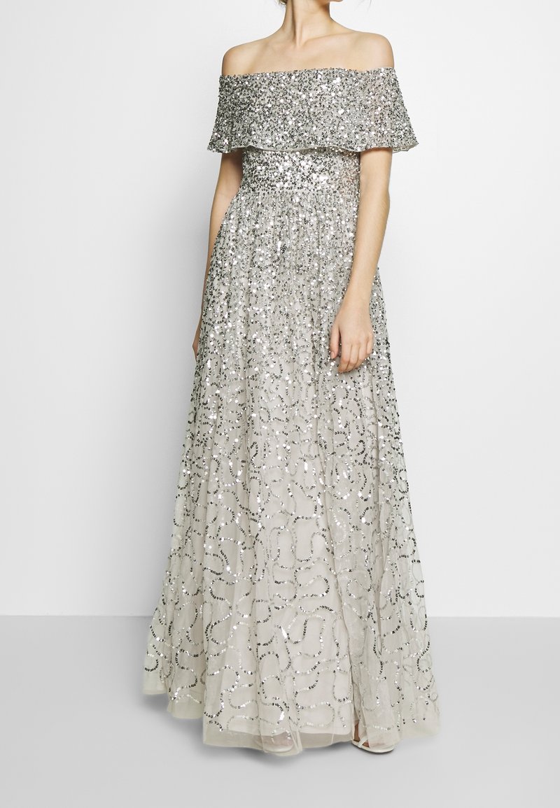 Femme portant une robe longue argentée à paillettes, sans bretelles, avec des motifs floraux et en spirale sur un fond clair.