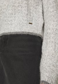 Hellgrauer Strickpullover mit strukturiertem Muster, mit geripptem Saum und dunkelgrauen Akzenten, kombiniert mit schwarzem Denim-Stoff.