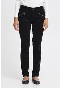 Fekete kordbársony nadrág karcsúsított szabással, szögletes cipzáros zsebekkel és gombos rögzítéssel. Fekete lapos cipővel viselve.