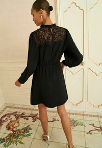 Črna obleka z čipkastim hrbtom, elastičnim pasom in nabranimi rokavi. Ima nabran ovratnik in odprtino v obliki ključavnice na hrbtu. Vidne srebrne sandale.