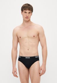Juodos trumpikės su baltu „BOSS" logotipu ant juosmens juostos. Audinys atrodo lygus ir prigludęs, pabrėžiantis paprastą dizainą.