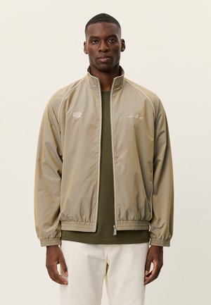 Giacca beige chiaro con zip, orli elastici, piping bianco a contrasto e logo ricamato sul petto, indossata sopra una camicia verde oliva.