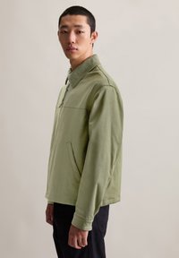 Helles grünes Jackett mit Kragen, Frontreißverschluss, Seitentaschen und glatter Textur. Getragen mit schwarzen Hosen vor einem neutralen Hintergrund.