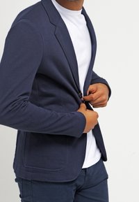 Man som knäpper en marinblå kavaj med en knapp som bärs över en vit t-shirt och mörka byxor, mot en plain vit bakgrund.