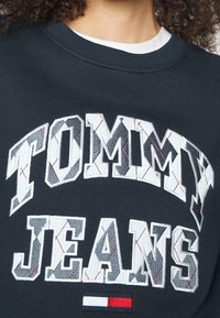 Tmavě modrá mikina s velkými bílými a vzorovanými písmeny "TOMMY JEANS". Obsahuje výšivky a malé logo ve spodní části.