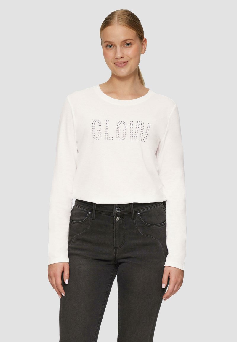 T-shirt blanc à manches longues avec des accents en cristal formant le mot "GLOW" sur le devant, associé à un jean foncé à taille haute. Texture en coton lisse.