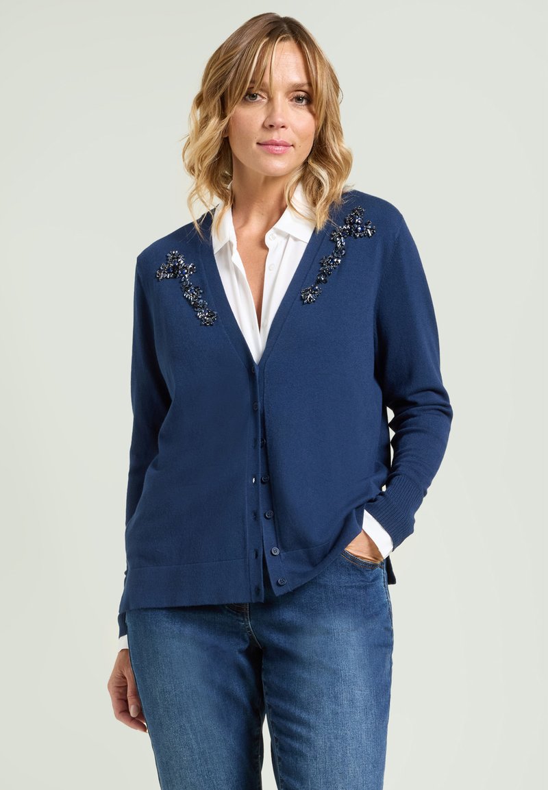 Marineblauwe cardigan van gebreid materiaal, met decoratieve versieringen op de schouders en een knoopsluiting aan de voorkant.