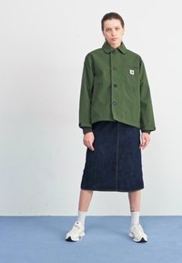 Veste verte courte avec une fermeture à bouton et une poche poitrine, associée à une jupe en denim foncé. Baskets blanches avec des accents noirs et des chaussettes blanches.
