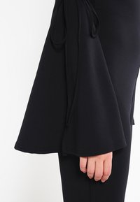 Svart klockärmad med justerbar knytdetalj, tillverkad av ett slätt tyg, som slutar vid mitten av underarmen, kombinerad med figurnära svarta kläder.