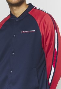 Giacca sportiva navy e rossa con chiusura a bottoni, logo sottile e accenti a righe a contrasto lungo le maniche. Tessuto morbido, design informale.