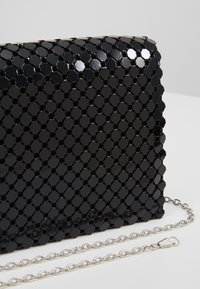 Musta kuusikulmainen paljettilaukku ketjuhihnalla, jossa on kiiltävä teksturoitu pinta ja geometrinen kuvio. Hopeiset yksityiskohdat näkyvät.