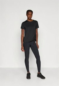 Ženska, oblečena v črno majico z gubami na strani, črne legice in črne športne copate, stoji pred enotno svetlo ozadje.