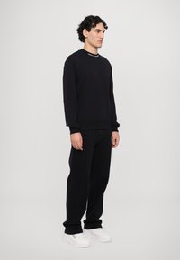 Felpa nera con colletto tondo e polsini a costine, abbinata a pantaloni neri e sneaker bianche. La felpa presenta del testo sul colletto.