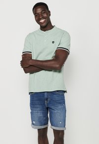 Polo shirt verde chiaro con maniche corte, caratterizzato da un colletto con bottoni e piccoli motivi. Abbinato a pantaloni corti in denim blu arrotolati ai polsini.