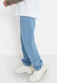 Jeans de mezclilla azul claro con un patrón de texto repetido "SIXTH JUNE", ajuste slim, combinados con zapatillas blancas gruesas. Textura suave, diseño casual.