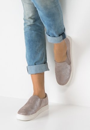 Person mit hellblauen hochgekrempelten Jeans und hellrosa Schlupfschuhen mit weißen Sohlen, steht mit einem Fuß an einer Wand.