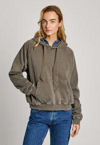 Sudadera gris con capucha, que presenta detalles de tachuelas en la parte frontal, un bolsillo delantero y puños de canalé. Lleva una camisa azul debajo.