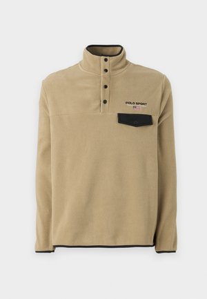 Beżowy pulower polarowy z czarnymi zatrzaskami, czarną lamówką, czarną kieszenią na zatrzask na piersi oraz logo "Polo Sport" z małą amerykańską flagą.