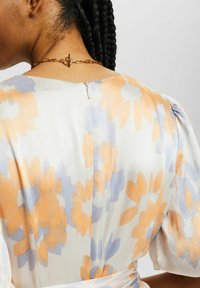 Robe fleurie en tissu doux de couleur blanche ornée de motifs orange et bleu, avec une fermeture éclair au dos et un détail en chaîne dorée au niveau du cou.