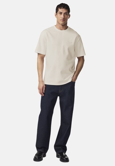 Mężczyzna ubrany w beżową koszulkę z krótkim rękawem, ciemnoniebieskie dżinsy i czarne mokasyny, stojący z jedną ręką w kieszeni na jednolitym tle.