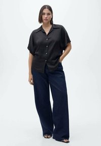Mørk oversized kortærmet skjorte med knapper og en blød tekstur, parret med brede, mørkeblå denimjeans og sorte sandaler med åbne tæer.