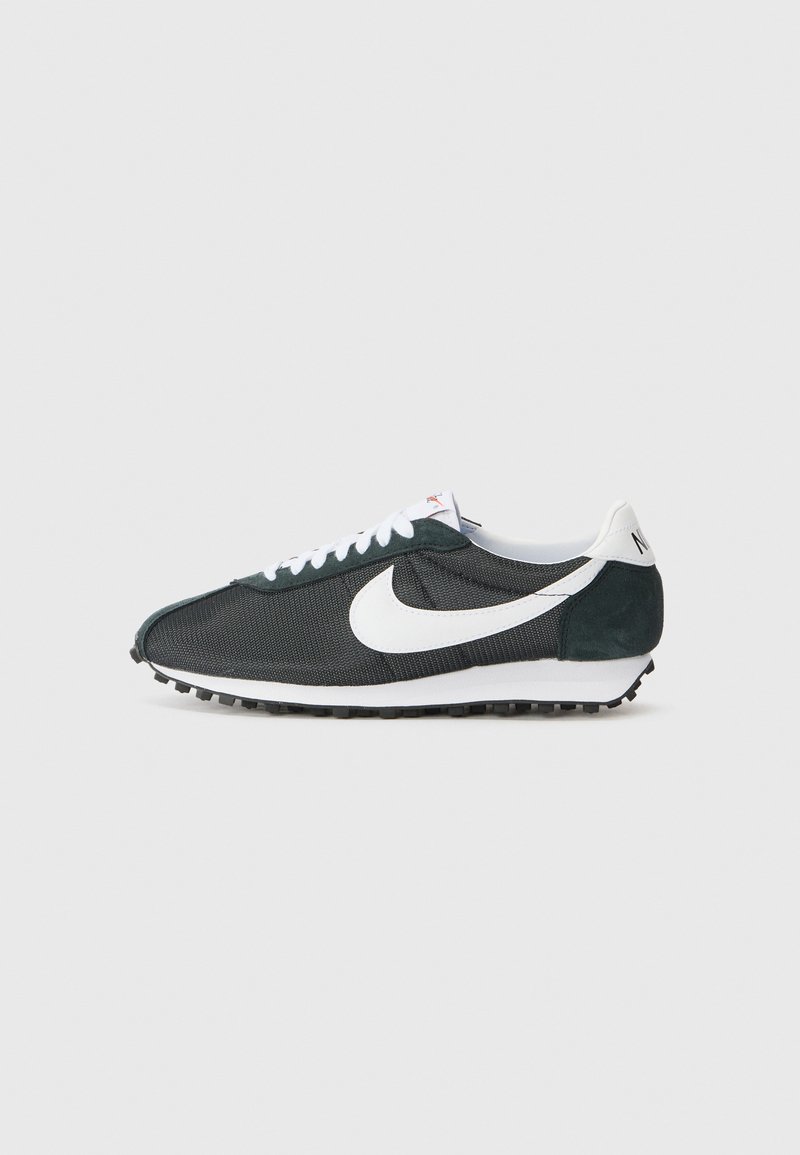 Zapatillas Nike de malla negra con swoosh blanco, cordones blancos y detalles en ante verde oscuro. Presenta una suela de goma negra texturizada.