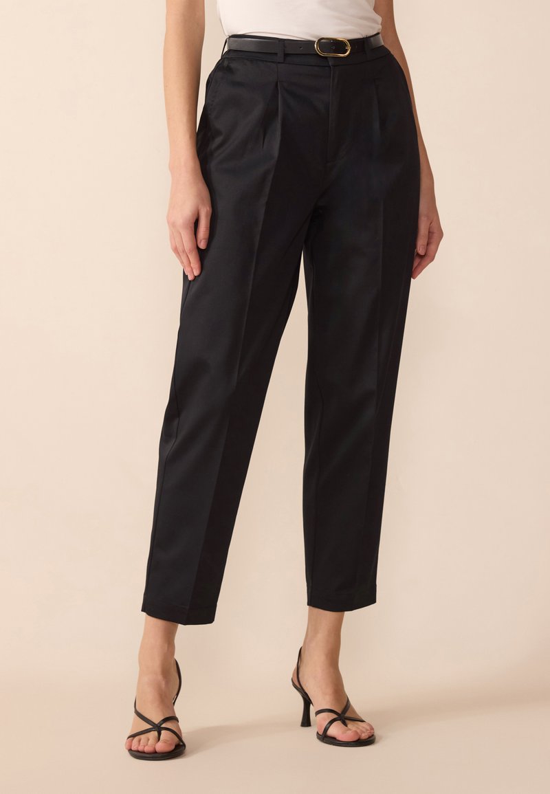 Black tailored trousers with a high waist, featuring pleats, smooth texture, and a tapered leg. Paired with black heeled sandals.