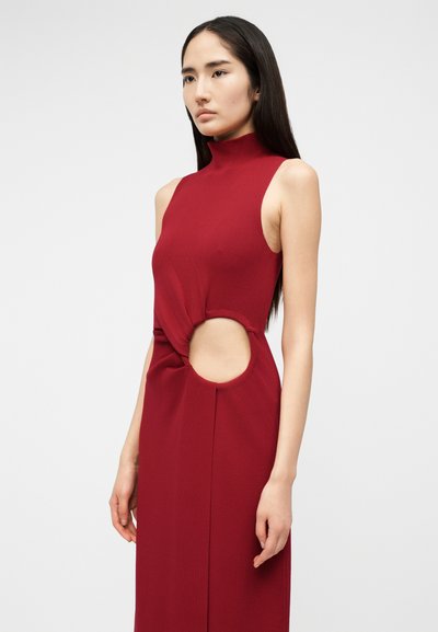 Hosszú sötét hajú nő, aki ujjatlan, magas nyakú piros ruhát visel, jobb oldalon a deréknál kör alakú kivágással.