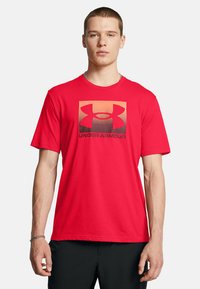 Camiseta de algodón roja con tejido transpirable, cuello redondo, mangas cortas y un logo de Under Armour en negro y naranja en la parte frontal.