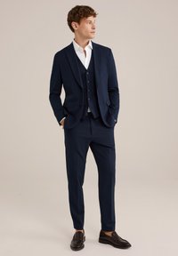 Costume trois pièces marine en tissu lisse, comprenant une veste ajustée, un gilet avec des boutons et un pantalon droit. Mocassins noirs.