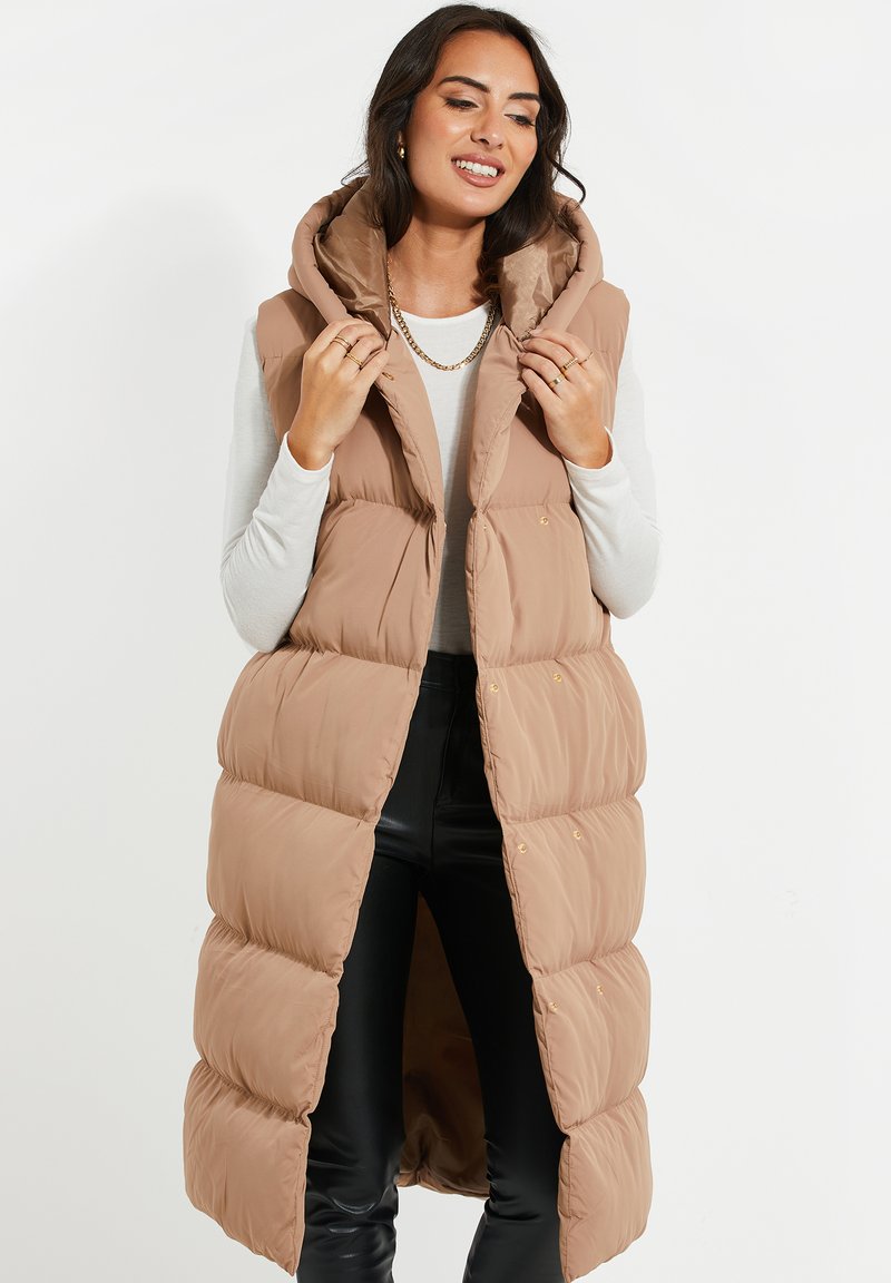 Gilet rembourré de couleur marron clair, longueur genou avec capuche, présentant une texture matelassée et des boutons-pression. Porté par-dessus un haut à manches longues.