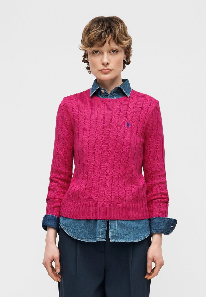 Jeune personne portant un pull en tricot torsadé rose vif par-dessus une chemise en jean boutonnée avec un pantalon sombre, faisant face à l'avant sur un fond uni.