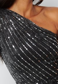 Robe asymétrique en tissu noir avec des sequins métalliques disposés en lignes verticales, ajoutant de la texture et des accents réfléchissants à la surface.