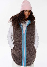 Hnědý, chlupatý vest s modrým a růžovým zipem, se bočními kapsami a vysokým límcem. Nosený přes bílou košili s dlouhým rukávem.