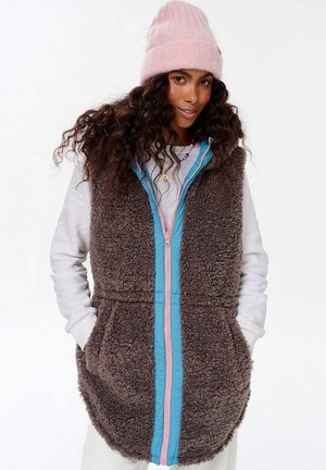 Brązowa, puszysta kamizelka z niebieskim i różowym suwakiem, z bocznymi kieszeniami i wysokim kołnierzem. Noszona na białej koszuli z długim rękawem.