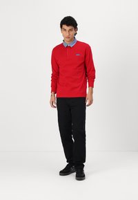 Red long-sleeve polo shirt with a blue collared design, featuring a buttoned neckline and logo detail. Paired with black pants and black shoes.