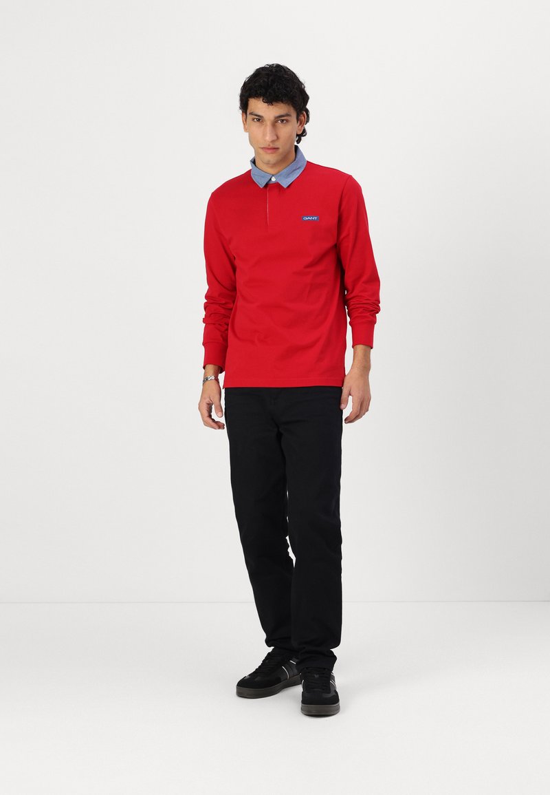 Rotes Langarm-Polohemd mit blauem Kragen-Design, ausgestattet mit einer geknöpften Ausschnittgestaltung und Logodetail. Kombiniert mit schwarzen Hosen und schwarzen Schuhen.