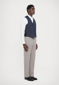 Gilet navy à cinq boutons, associé à un pantalon gris clair. La chemise est blanche avec une texture subtile. Des chaussures noires complètent le look.