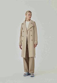 Woman wearing beige double-breasted trench coat, matching wide-leg pants, white top, and light blue platform sandals standing against plain background.