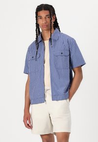 Herren Kurzarmhemd im karrierten Muster in Marineblau und Weiß, mit zwei Brusttaschen und kragenlöse Ausschnitt. Kombiniert mit beigen Shorts.