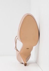 Chaussure à talon haut beige avec une semelle pointue et une bride à la cheville, photographiée depuis le bord inférieur sur un fond blanc en coin.