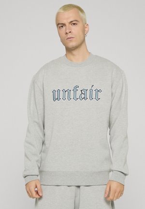Uomo biondo con orecchini che indossa una felpa e pantaloni grigio chiaro con la parola "unfair" in carattere Old English nero sul petto.