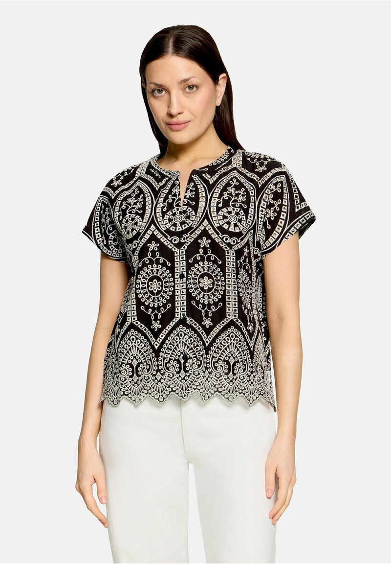 Femme aux cheveux lisses et foncés, portant un haut noir avec une broderie géométrique blanche complexe et un pantalon blanc, debout devant un fond clair.