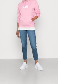Persona con una felpa rosa di Levi's, jeans blu e sneaker platform bianche in piedi davanti a uno sfondo bianco semplice.